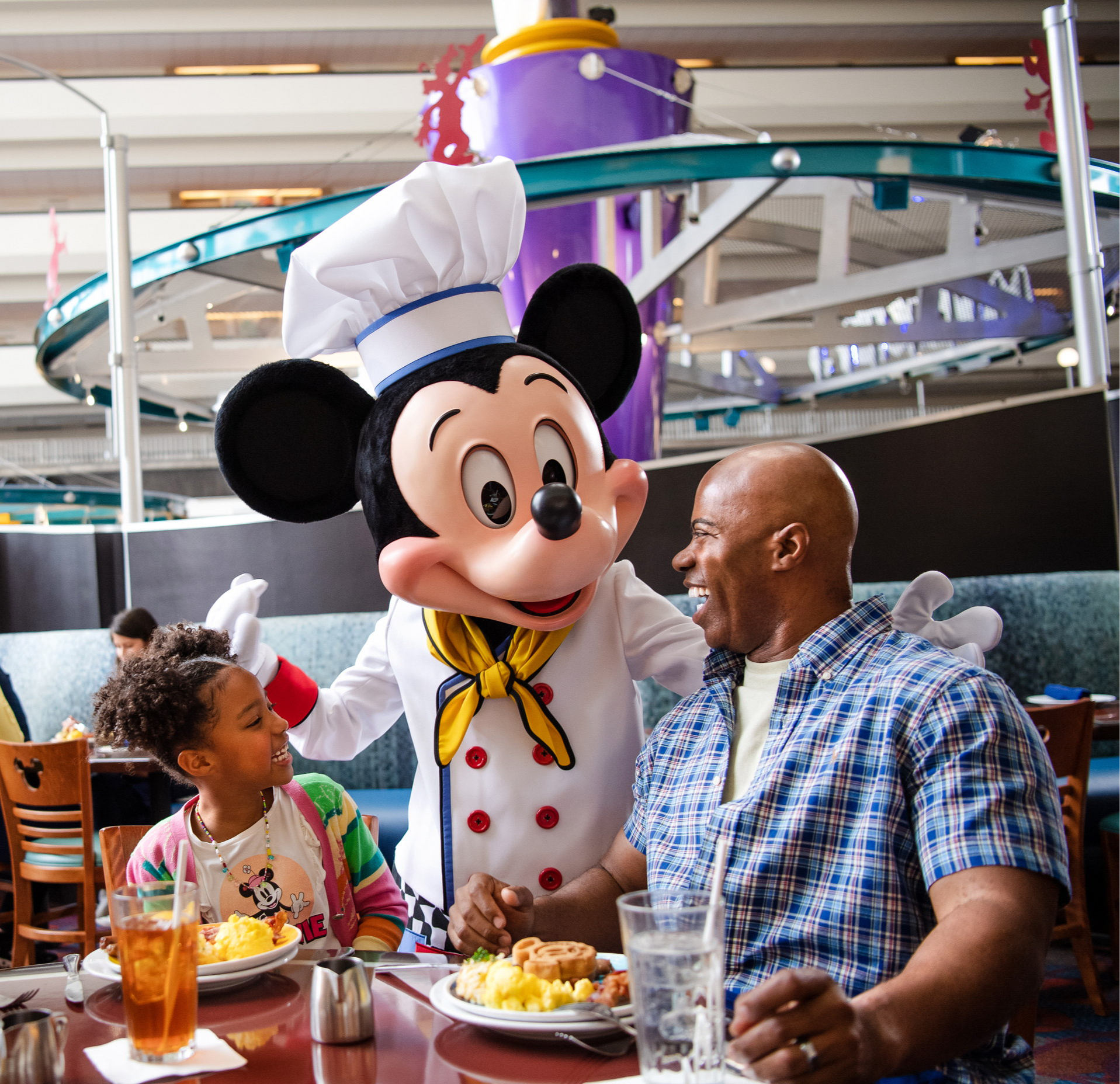 Mickey en toque de chef salue une fillette et son père attablés devant leur assiette, sous le rail du monorail, au Chef Mickey’s Mickey en toque de chef salue une fillette et son père attablés devant leur assiette, sous le rail du monorail, au Chef Mickey’s